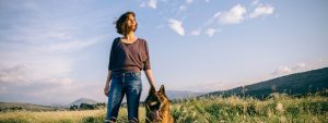 Woman with a dog in a field looking toward the sun on clear day.